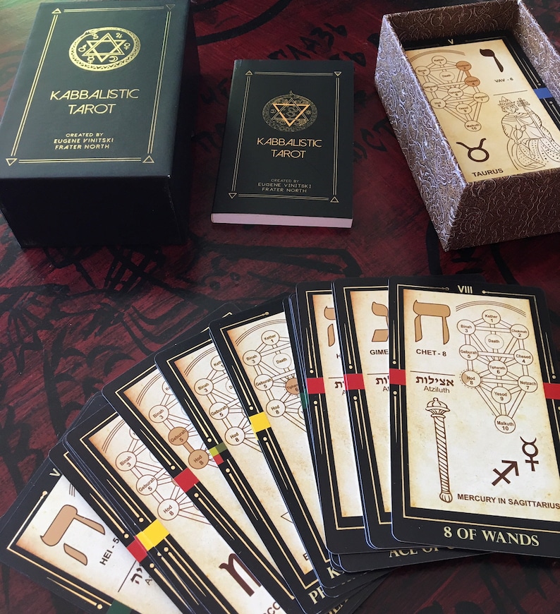 May include: A spread of black and gold tarot cards with Hebrew letters and illustrations of trees. The cards are laid out on a dark red wooden surface. The cards are from the Kabbalistic Tarot deck.