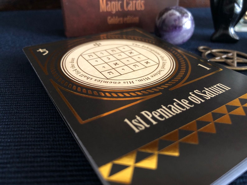 May include: A black and gold card with the text "1st Pentacle of Saturn" and a gold circle with Hebrew characters and the text "His enemies shall lick the dust.  Kneel before Him."