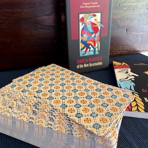 Peut inclure: Un jeu de cartes de Tarot de Marseille avec un motif g&eacute;om&eacute;trique jaune et bleu au dos. La bo&icirc;te des cartes est noire avec du texte rouge qui dit "Tarot de Marseille de la Nouvelle Incarnation".