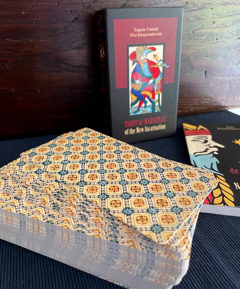 Peut inclure: Un jeu de cartes de Tarot de Marseille avec un motif g&eacute;om&eacute;trique jaune et bleu au dos. La bo&icirc;te des cartes est noire avec du texte rouge qui dit "Tarot de Marseille de la Nouvelle Incarnation".