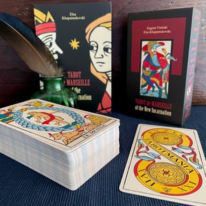Peut inclure: Un jeu de cartes de Tarot de Marseille avec une bo&icirc;te noire sur laquelle est inscrit "Tarot de Marseille de la Nouvelle Incarnation" ainsi que les noms "Eug&egrave;ne Vinitski" et "Elsa Khapatnukovski". Le jeu est empil&eacute; sur une surface bleue avec une seule carte face visible, montrant la carte "Le Monde" avec un design jaune et rouge.