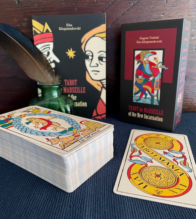 Peut inclure: Un jeu de cartes de Tarot de Marseille avec une bo&icirc;te noire sur laquelle est inscrit "Tarot de Marseille de la Nouvelle Incarnation" ainsi que les noms "Eug&egrave;ne Vinitski" et "Elsa Khapatnukovski". Le jeu est empil&eacute; sur une surface bleue avec une seule carte face visible, montrant la carte "Le Monde" avec un design jaune et rouge.