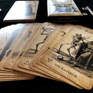 May include: A deck of tarot cards with vintage-style illustrations, featuring figures like "The Emperor" and "King." The cards are fanned out, with a box and keys in the background. The cards have a sepia tone.