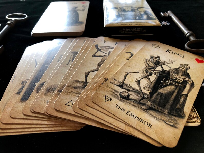 May include: A deck of tarot cards with vintage-style illustrations, featuring figures like "The Emperor" and "King." The cards are fanned out, with a box and keys in the background. The cards have a sepia tone.