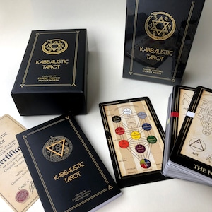May include: A black box with gold lettering that reads "Kabbalistic Tarot" and a gold star of David symbol. The box is open and the tarot cards are visible. The cards are black with gold lettering and illustrations. The cards are arranged in a fan shape. The text on the cards reads "Kabbalistic Tarot" and "The Fool".