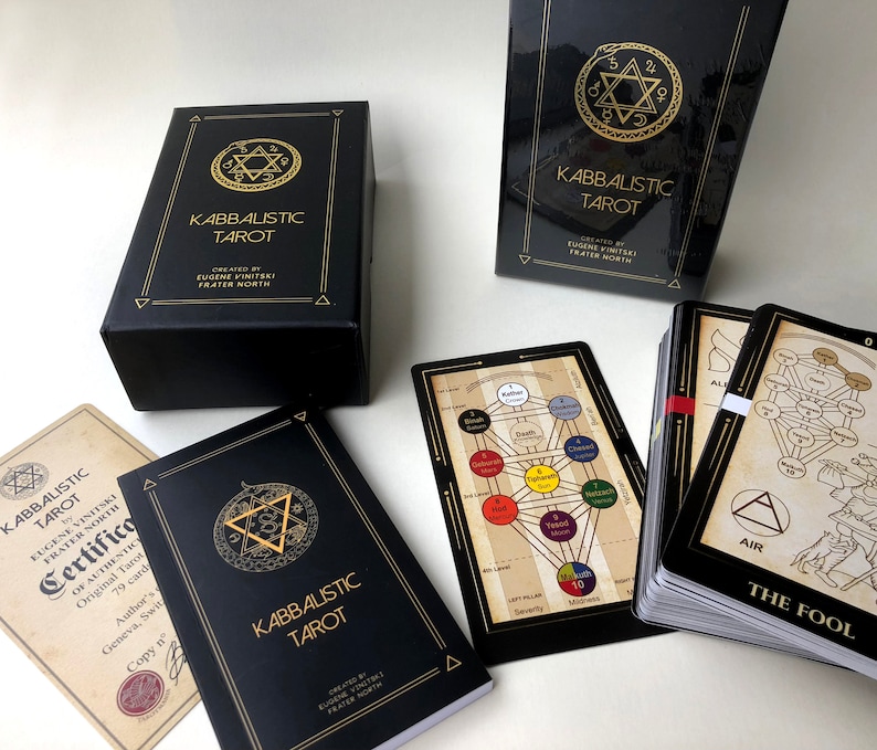 May include: A black box with gold lettering that reads "Kabbalistic Tarot" and a gold star of David symbol. The box is open and the tarot cards are visible. The cards are black with gold lettering and illustrations. The cards are arranged in a fan shape. The text on the cards reads "Kabbalistic Tarot" and "The Fool".