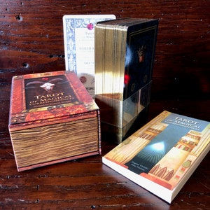 May include: A set of Tarot of Magical Correspondences cards in a brown box with a gold trim. The box has a skull and horns on the front. The cards are wrapped in gold foil and have a blue and gold design. The book is titled "Tarot of Magical Correspondences" and has a gold and white design.