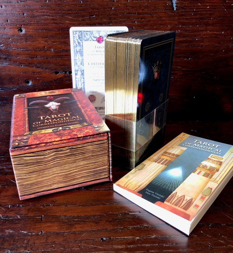 May include: A set of Tarot of Magical Correspondences cards in a brown box with a gold trim. The box has a skull and horns on the front. The cards are wrapped in gold foil and have a blue and gold design. The book is titled "Tarot of Magical Correspondences" and has a gold and white design.