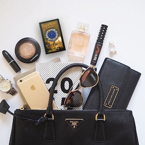 May include: A black leather handbag with gold hardware and a gold Prada logo. The bag is open and filled with various items, including a phone, sunglasses, a tarot deck, makeup, and a notebook.