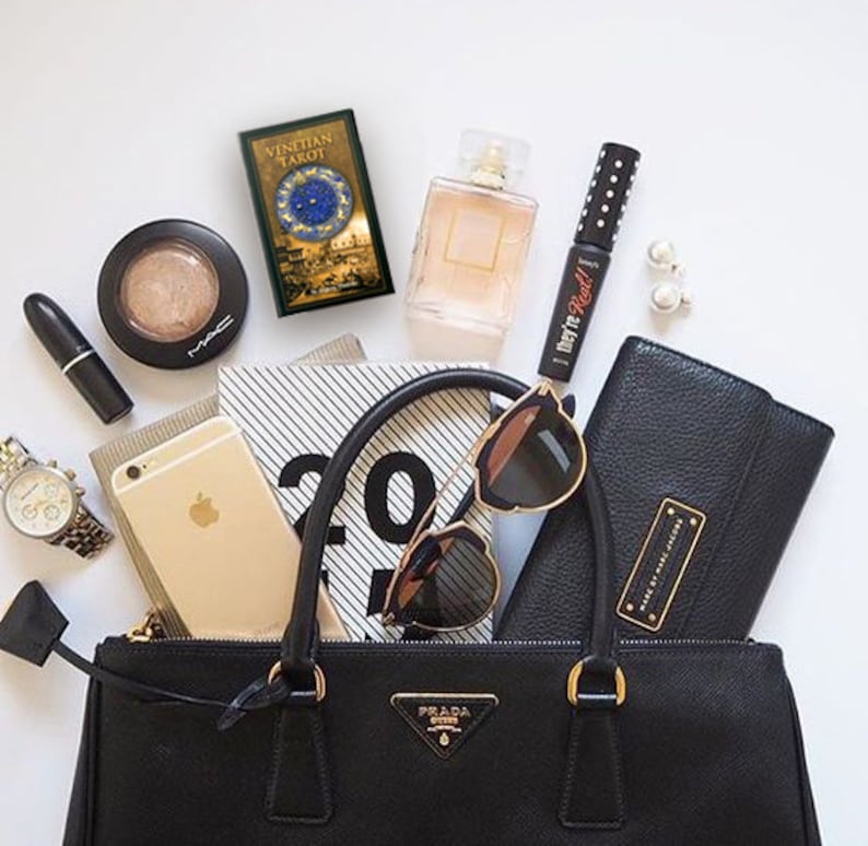 May include: A black leather handbag with gold hardware and a gold Prada logo. The bag is open and filled with various items, including a phone, sunglasses, a tarot deck, makeup, and a notebook.