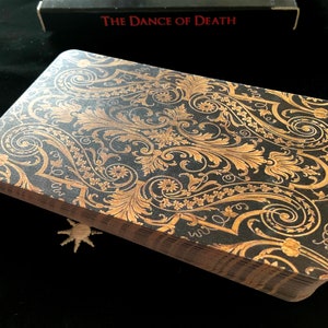 May include: A deck of tarot cards with a black background and gold floral design. The cards are stacked, showing the detailed pattern on the top card. The box in the background reads "The Dance of Death".