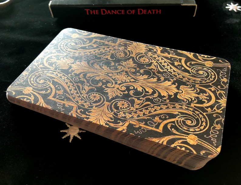May include: A deck of tarot cards with a black background and gold floral design. The cards are stacked, showing the detailed pattern on the top card. The box in the background reads "The Dance of Death".