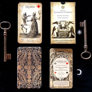 May include: An oracle card deck titled "The Dance of Death" with illustrations and text. The deck includes cards, a box, and a certificate of authenticity. Two antique keys and decorative celestial shapes are also present.