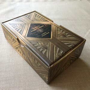 May include: A gold box with an art deco design. The box is closed and has a label that reads "Designed by Eugene Vinidzal, Geneva, Switzerland".