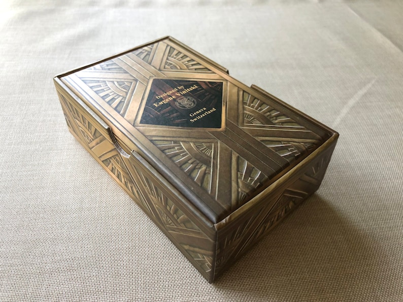 May include: A gold box with an art deco design. The box is closed and has a label that reads "Designed by Eugene Vinidzal, Geneva, Switzerland".