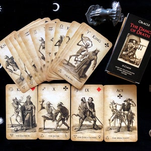May include: An oracle card deck titled "The Dance of Death" with 33 cards, featuring vintage-style illustrations of skeletons and figures. The cards are fanned out on a black surface, accompanied by a small box, two antique keys, and a decorative metal hand.