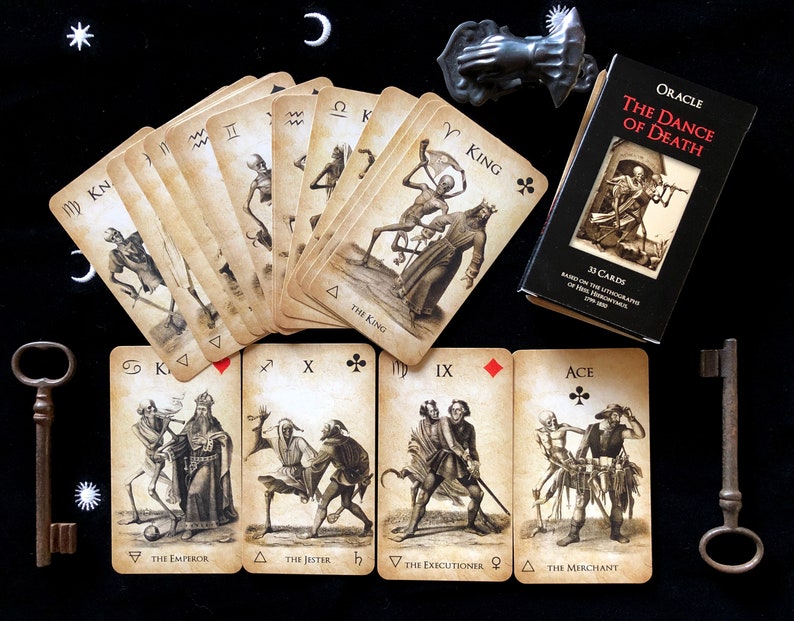 May include: An oracle card deck titled "The Dance of Death" with 33 cards, featuring vintage-style illustrations of skeletons and figures. The cards are fanned out on a black surface, accompanied by a small box, two antique keys, and a decorative metal hand.