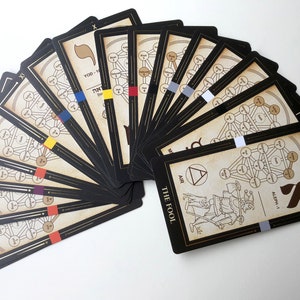 May include: A spread of 22 tarot cards with black borders and gold accents. The cards feature a tree of life design with Hebrew letters and a color-coded system. The card in the front is The Fool, with a depiction of a man walking with a staff and a backpack. The card is labeled with the Hebrew letter Aleph-1 and the number 0.