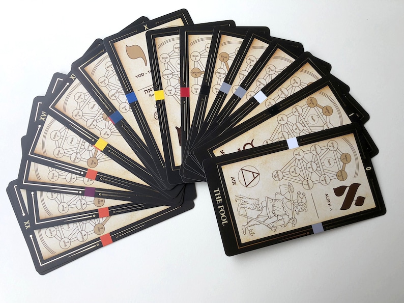 May include: A spread of 22 tarot cards with black borders and gold accents. The cards feature a tree of life design with Hebrew letters and a color-coded system. The card in the front is The Fool, with a depiction of a man walking with a staff and a backpack. The card is labeled with the Hebrew letter Aleph-1 and the number 0.