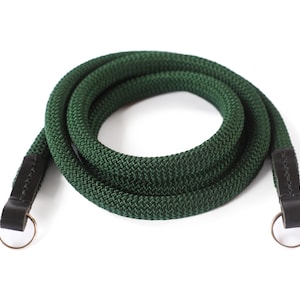 Può includere: Una tracolla per fotocamera in corda verde scuro con estremità in pelle nera e anelli metallici. La corda è arrotolata su uno sfondo bianco. La tracolla è progettata per essere attaccata a una fotocamera.