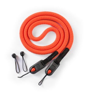 Puede incluir: Correa de cámara naranja brillante hecha de una cuerda gruesa y tejida. La correa tiene conectores negros con botones rojos y dos sujetadores de goma negros. La correa está enrollada sobre un fondo blanco.