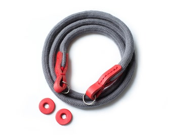 May include: A gray camera strap with red leather accents and two red metal rings.