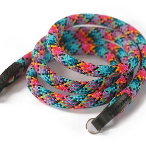 Op de afbeelding: Een kleurrijke camerariem met een geweven patroon in roze, blauw, geel en zwart. De riem heeft zwarte leren uiteinden met metalen ringen. De riem is opgerold op een witte achtergrond.