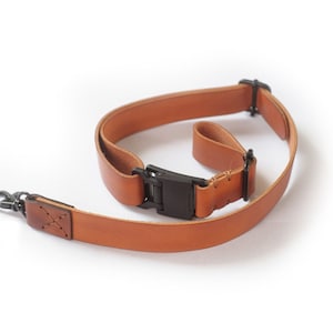 May include: A brown leather camera strap with a black adjustable buckle and a metal hook. The strap is designed for carrying a camera around the neck or shoulder.