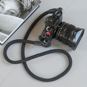May include: A black camera with a black lens and a black camera strap. The camera has a red button on the top.