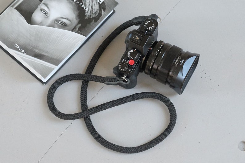 May include: A black camera with a black lens and a black camera strap. The camera has a red button on the top.