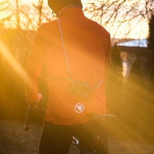 May include: A person wearing a bright orange cycling jacket with a white logo on the back, a black helmet, and black cycling trousers, riding a bicycle on a dirt path at sunset.