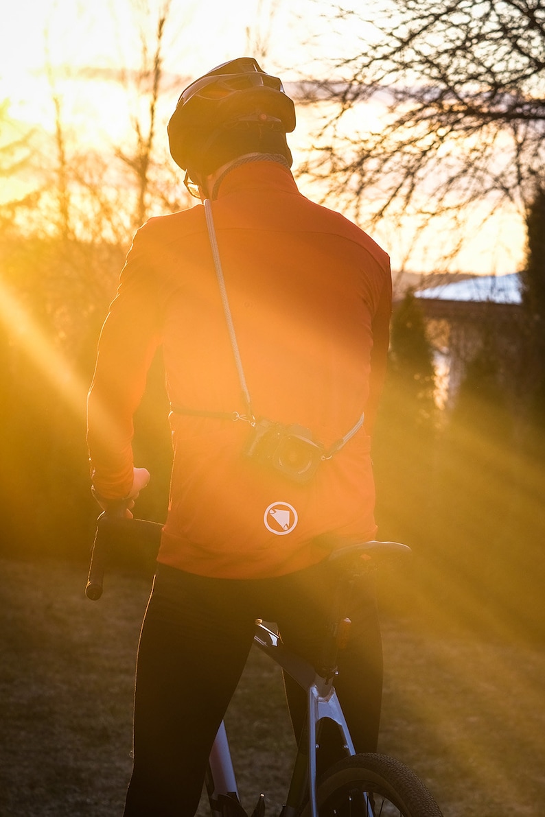 May include: A person wearing a bright orange cycling jacket with a white logo on the back, a black helmet, and black cycling trousers, riding a bicycle on a dirt path at sunset.