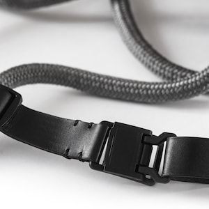 May include: A black leather camera strap with a grey rope and a black plastic buckle.
