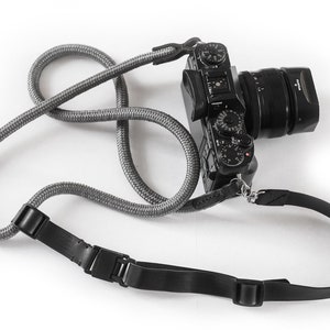 May include: A black camera with a lens attached is hanging from a grey and black camera strap. The strap has two adjustable buckles.