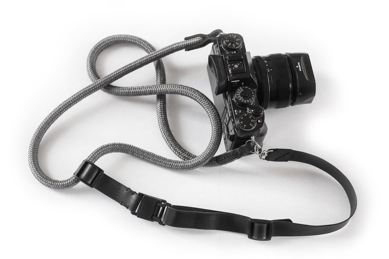 May include: A black camera with a lens attached is hanging from a grey and black camera strap. The strap has two adjustable buckles.