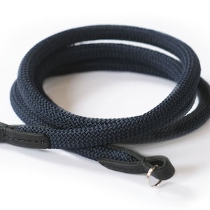 Puede incluir: Una correa de cámara azul oscuro hecha de tejido tejido con extremos de cuero negro y un anillo de metal plateado.