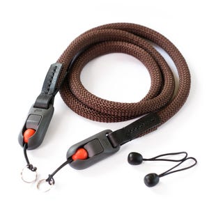 May include: A brown camera strap with a braided rope design. The strap features black leather accents, gray plastic adjustment buckles with orange buttons, and small black and silver attachments. The strap is designed for a camera.