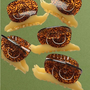 May include: Four hair claw clips shaped like snails. Each clip features a cream-coloured base with a brown and black tortoiseshell pattern on the shell portion. The snail-shaped clips have a spring mechanism and teeth for securing hair in place.