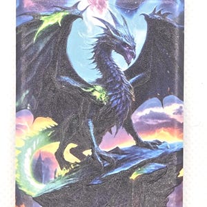 Pode incluir: Um íman retangular com uma ilustração detalhada de um dragão escuro alado pousado numa formação rochosa irregular. O dragão tem detalhes em verde brilhante nas asas e na cauda, contra um céu com nuvens roxas e cor de laranja.
