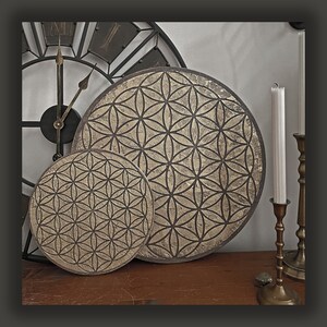 May include: Two gold-toned decorative wall hangings with a geometric pattern resembling a flower. The larger hanging is round and the smaller hanging is slightly smaller and round. The hangings are on a wooden surface.