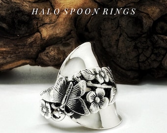 Op de afbeelding: Een zilveren lepelring met een vlinder- en bloemenontwerp. De ring heeft een gedetailleerde vlinder en bloemen, met de tekst "HALO SPOON RINGS" boven de ring. De ring staat tegen een houten achtergrond.