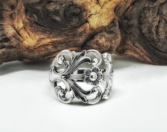 Op de afbeelding: Zilveren lepelring met ingewikkelde bloemen- en krulontwerpen. De ring heeft een centrale bloem en de tekst "HALO SPOON RINGS". De ring staat tegen een witte achtergrond met een houten achtergrond.