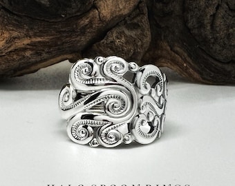 Op de afbeelding: Een zilveren ring met een zwierig, versierd ontwerp. De ring is breed en heeft een getextureerd oppervlak. HALO SPOON RINGS staat in zwarte tekst onder de ring.