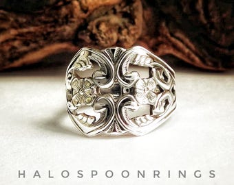 Op de afbeelding: Een zilveren ring met een sierlijk bloemmotief. De ring heeft opengewerkte details met bloemen en krullende patronen. De tekst "HALOSPOONRINGS" staat onderaan de afbeelding. De ring staat tegen een neutrale achtergrond.
