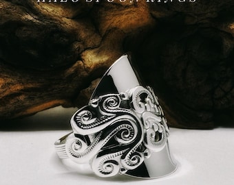 Op de afbeelding: Een zilverkleurige lepelring met een gedetailleerd, verhoogd krulpatroon op de voorzijde. Het metaal is gepolijst en reflecterend, met de tekst HALO SPOON RINGS in witte letters boven de ring weergegeven.