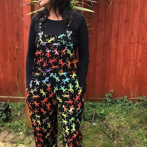 May include: Black overalls with a rainbow lizard print. The overalls have adjustable straps and pockets.