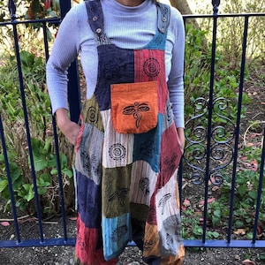 May include: A colourful patchwork jumpsuit with a variety of patterns and colours, including red, blue, and green. The jumpsuit has a front pocket with an eye design and black floral patterns. The loose-fitting trousers end at the ankles.