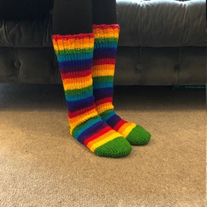 Op de afbeelding: Kniehoge sokken met een levendig regenboogpatroon. De sokken hebben horizontale strepen in rood, oranje, geel, groen, blauw en paars. De tenen zijn groen en de manchetten rood.