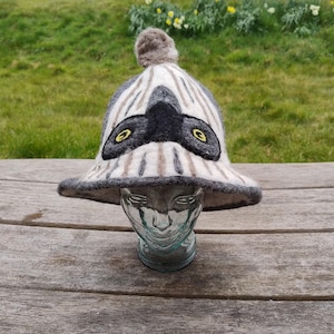 May include: A gray and white striped felt hat with a black felt owl design and yellow felt eyes. The hat has a rounded crown and a wide brim.