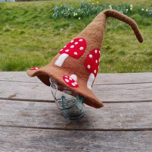 Puede incluir: Un sombrero de bruja de fieltro marrón con una punta larga y puntiaguda y un ala ancha. El sombrero está decorado con setas de fieltro rojo y blanco.